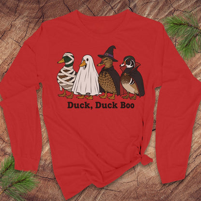 A red long-sleeve t-shirt with a graphic of four ducks in Halloween costumes (mummy, ghost, witch, and wood duck) and the text 'Duck, Duck Boo' printed below, displayed flat on a rustic wood background with pine needles.