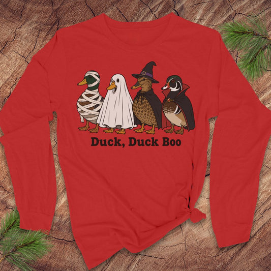 A red long-sleeve t-shirt with a graphic of four ducks in Halloween costumes (mummy, ghost, witch, and wood duck) and the text 'Duck, Duck Boo' printed below, displayed flat on a rustic wood background with pine needles.