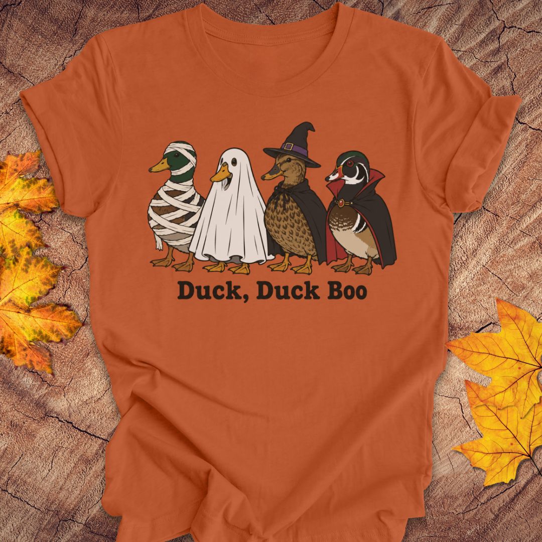 An orange short-sleeve t-shirt with a graphic of four cartoon ducks in Halloween costumes (mummy, ghost, witch, and wood duck) and the text 'Duck, Duck Boo' printed below. The shirt is displayed flat on a rustic wood background decorated with autumn leaves.