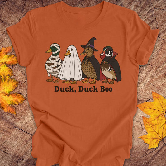 An orange short-sleeve t-shirt with a graphic of four cartoon ducks in Halloween costumes (mummy, ghost, witch, and wood duck) and the text 'Duck, Duck Boo' printed below. The shirt is displayed flat on a rustic wood background decorated with autumn leaves.