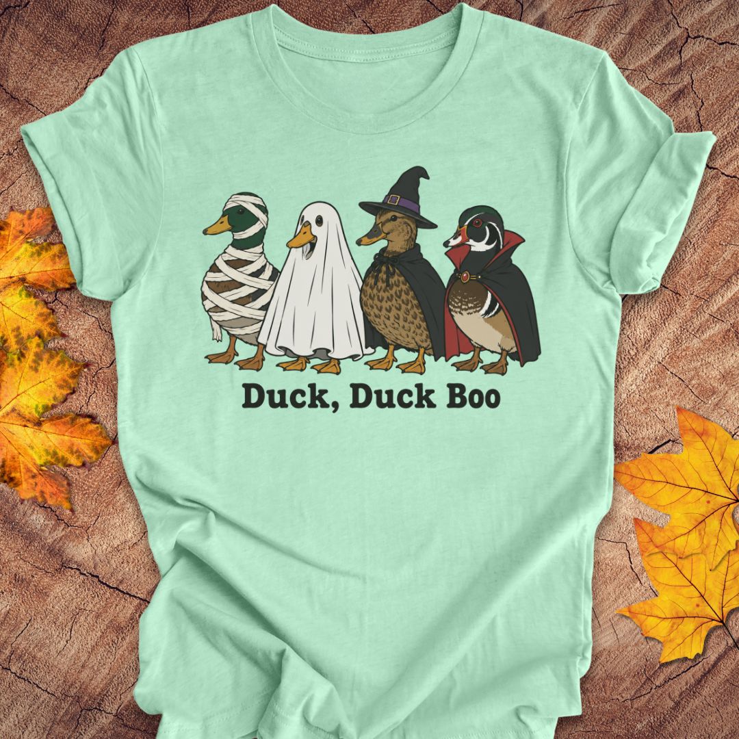 A mint green short-sleeve t-shirt laid flat on a rustic wood background with autumn leaves. The shirt features a graphic of four cartoon ducks dressed in Halloween costumes (mummy, ghost, witch, and wood duck) with the text 'Duck, Duck Boo' printed below.