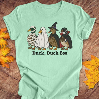 A mint green short-sleeve t-shirt laid flat on a rustic wood background with autumn leaves. The shirt features a graphic of four cartoon ducks dressed in Halloween costumes (mummy, ghost, witch, and wood duck) with the text 'Duck, Duck Boo' printed below.