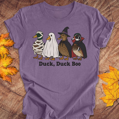 A purple short-sleeve t-shirt laid flat on a rustic wood background with autumn leaves. The shirt features a graphic of four ducks dressed in Halloween costumes (mummy, ghost, witch, and wood duck) with the text 'Duck, Duck Boo' printed below.