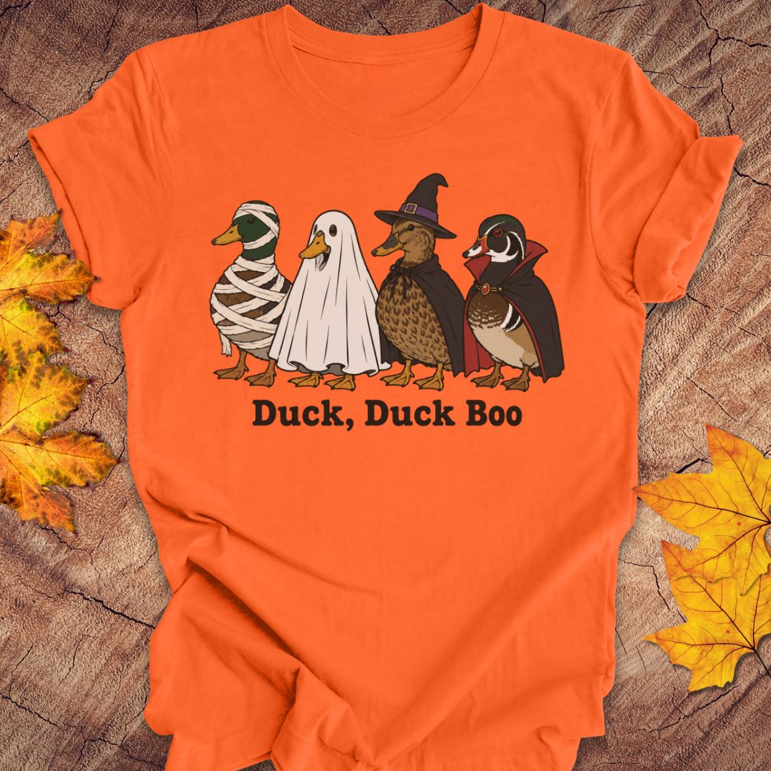 An orange short-sleeve t-shirt laid flat on a rustic wood surface with autumn leaves around it. The shirt features a graphic of four cartoon ducks dressed in Halloween costumes with the text 'Duck, Duck Boo' underneath.