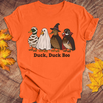 An orange short-sleeve t-shirt laid flat on a rustic wood surface with autumn leaves around it. The shirt features a graphic of four cartoon ducks dressed in Halloween costumes with the text 'Duck, Duck Boo' underneath.
