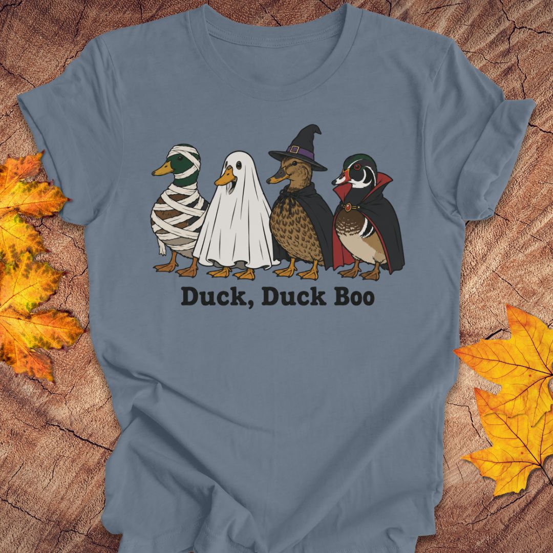 A slate blue crewneck t-shirt featuring a graphic of four duck characters in Halloween costumes (mummy, ghost, witch, and woodpecker) with the text 'Duck, Duck Boo' underneath, displayed on a rustic wood background with autumn leaves.