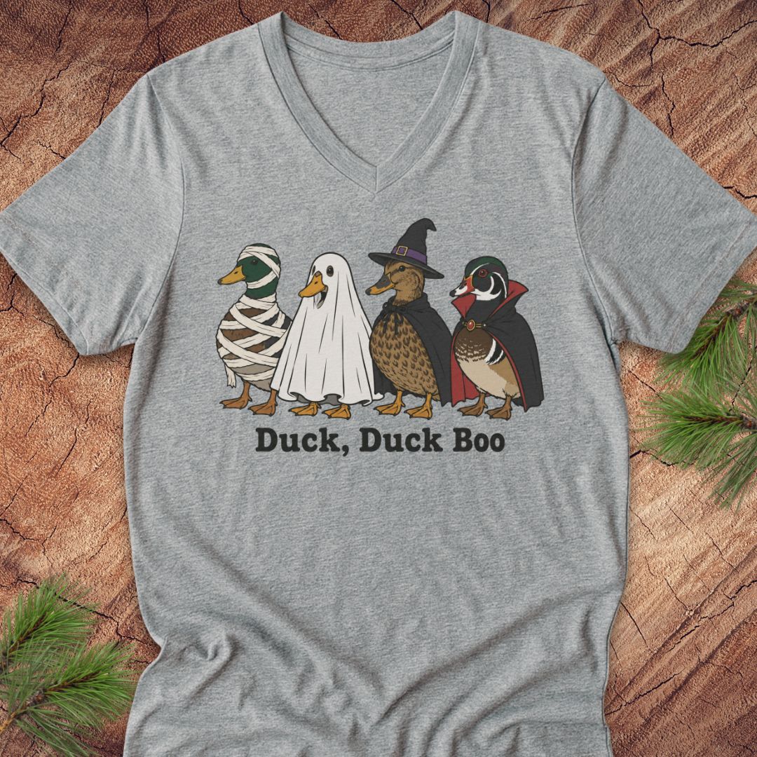 A heather gray V-neck t-shirt featuring a graphic of four cartoon ducks in Halloween costumes (mummy, ghost, witch, and vampire) with the text 'Duck, Duck Boo' printed below. The shirt is laid flat on a rustic wood background with pine branches in the corners.