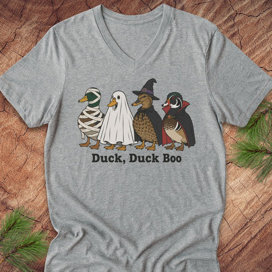 A heather gray V-neck t-shirt featuring a graphic of four cartoon ducks in Halloween costumes (mummy, ghost, witch, and vampire) with the text 'Duck, Duck Boo' printed below. The shirt is laid flat on a rustic wood background with pine branches in the corners.