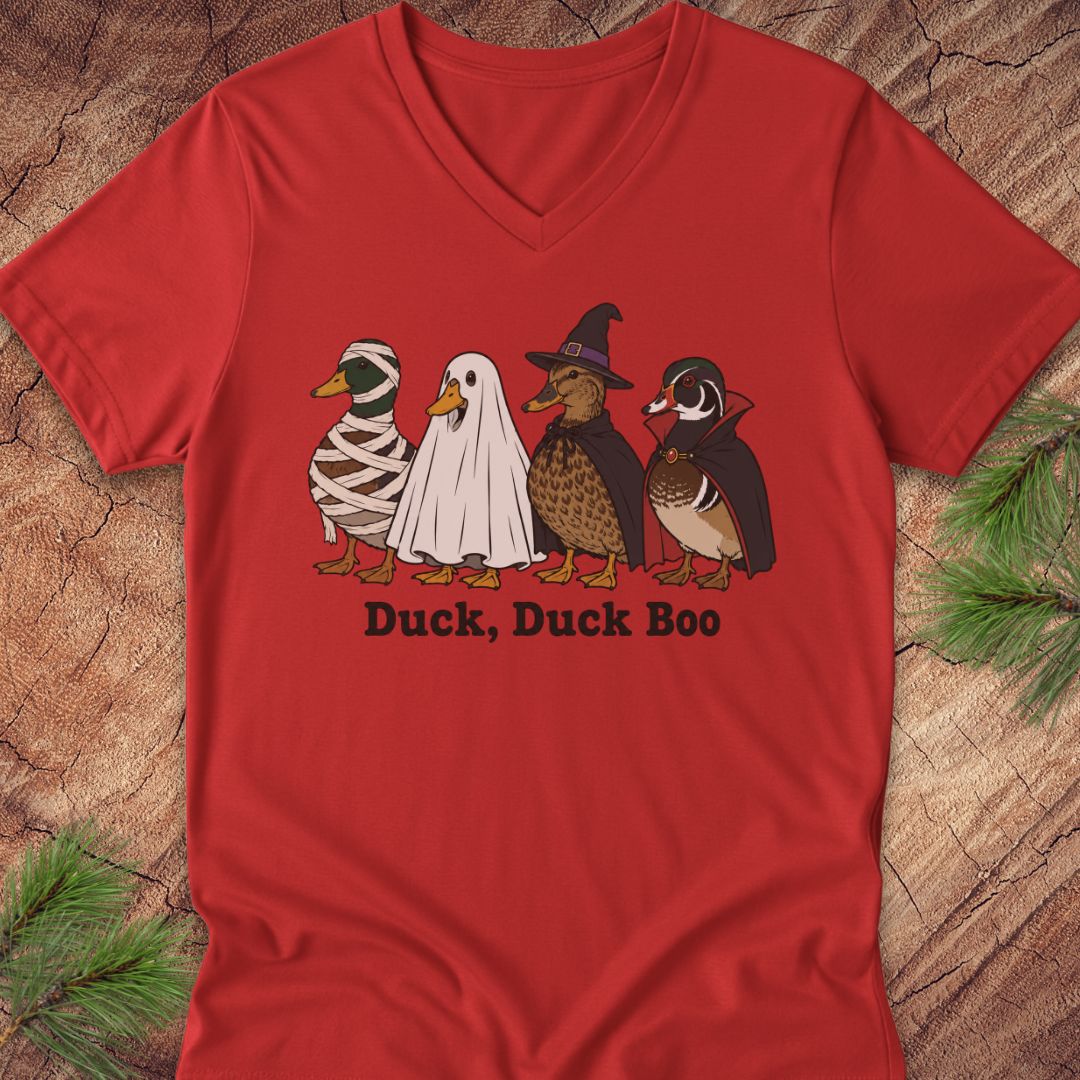 A red V-neck t-shirt featuring a graphic of four ducks in Halloween costumes (mummy, ghost, witch, and normal duck) with the text 'Duck, Duck Boo' underneath, displayed flat on a rustic wood background with pine branches.