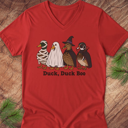 A red V-neck t-shirt featuring a graphic of four ducks in Halloween costumes (mummy, ghost, witch, and normal duck) with the text 'Duck, Duck Boo' underneath, displayed flat on a rustic wood background with pine branches.