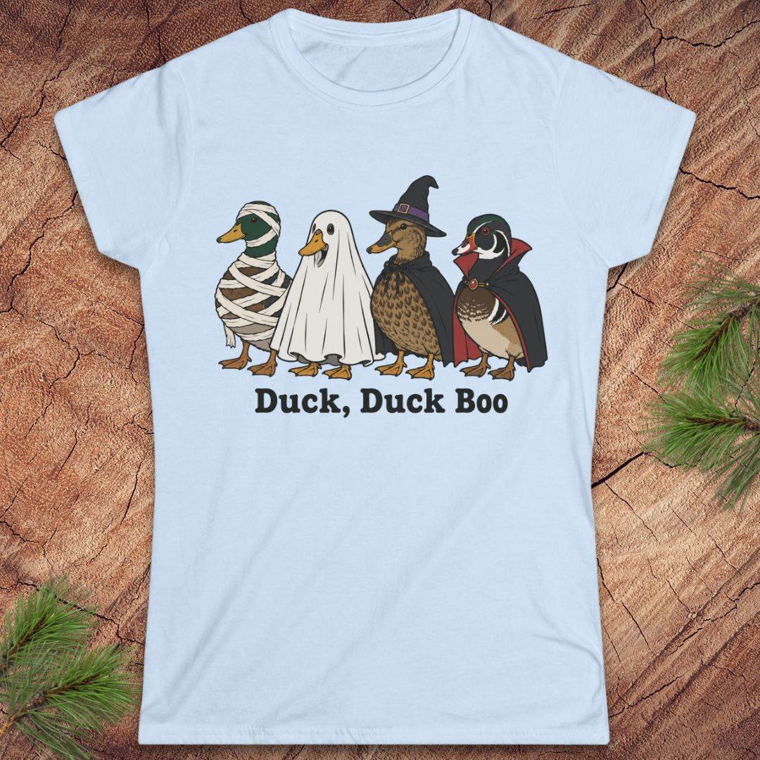 A light blue short-sleeve t-shirt with a graphic of four ducks in Halloween costumes (mummy, ghost, witch, and wood duck) and the text 'Duck, Duck Boo' printed below it. The shirt is displayed flat on a rustic wood background with pine needles in the corners.