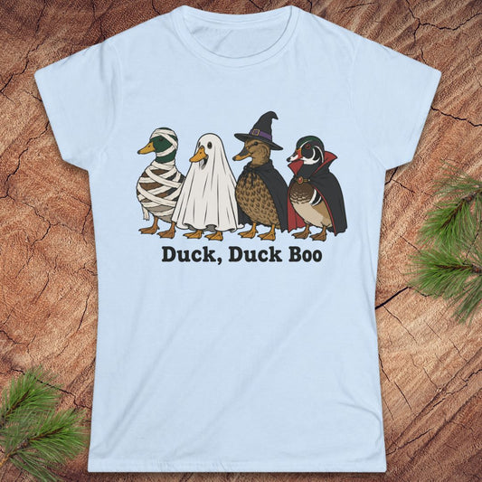 A light blue short-sleeve t-shirt with a graphic of four ducks in Halloween costumes (mummy, ghost, witch, and wood duck) and the text 'Duck, Duck Boo' printed below it. The shirt is displayed flat on a rustic wood background with pine needles in the corners.