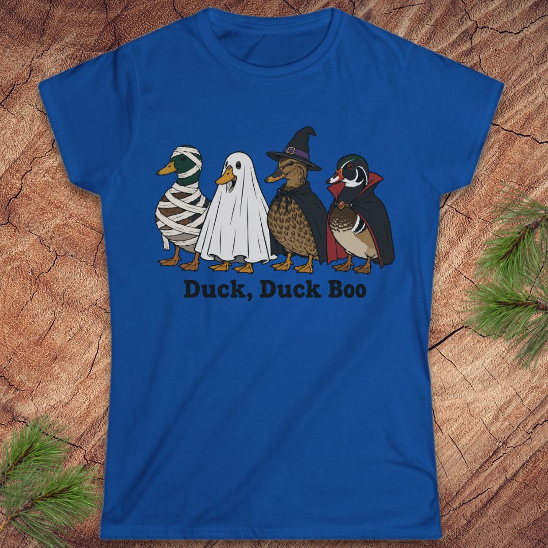 A royal blue short-sleeve t-shirt with a graphic of four duck characters in Halloween costumes (mummy, ghost, witch, and wood duck) and the text 'Duck, Duck Boo' printed below. The shirt is displayed flat on a rustic wood background with pine needles in the corners.