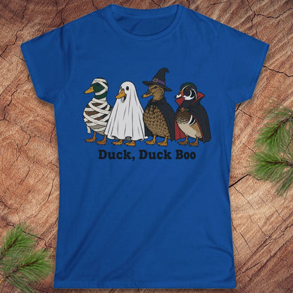 A royal blue short-sleeve t-shirt with a graphic of four duck characters in Halloween costumes (mummy, ghost, witch, and wood duck) and the text 'Duck, Duck Boo' printed below. The shirt is displayed flat on a rustic wood background with pine needles in the corners.
