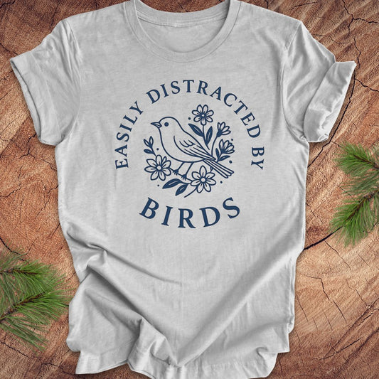 Gray t-shirt with a bird graphic and text 'easily distracted by birds' on a wood background.