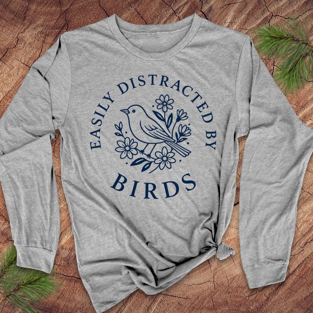 A heather gray long-sleeve t-shirt with a navy blue graphic print of a bird and flowers, laid flat on a rustic wood background with pine needles.