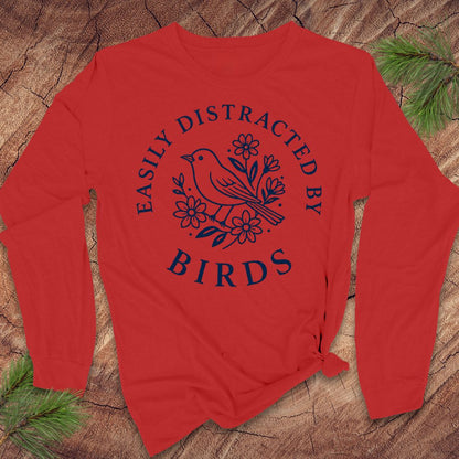 A red long-sleeve t-shirt with a navy blue graphic print of a bird and flowers, laid flat on a rustic wood background with pine branches.