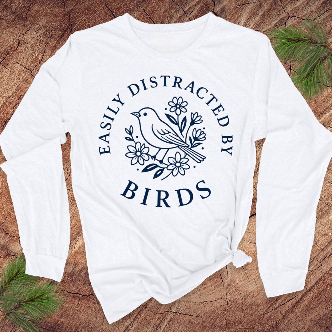 A white long-sleeve t-shirt with a navy blue graphic print of a bird and flowers, laid flat on a rustic wood background with pine branches.