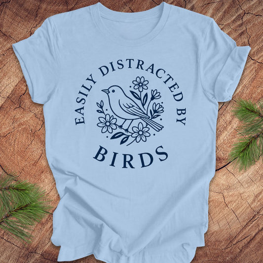 A light blue short-sleeve t-shirt with a dark navy blue graphic print featuring a bird and flowers, with the text 'EASILY DISTRACTED BY BIRDS' arched above and below the design. The t-shirt is laid flat on a rustic wood background with pine branches in the corners.