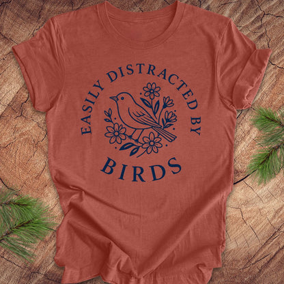 A terracotta-colored short-sleeve t-shirt with a navy blue graphic print reading 'EASILY DISTRACTED BY BIRDS' around a bird and floral design, laid flat on a rustic wood background with pine branches.