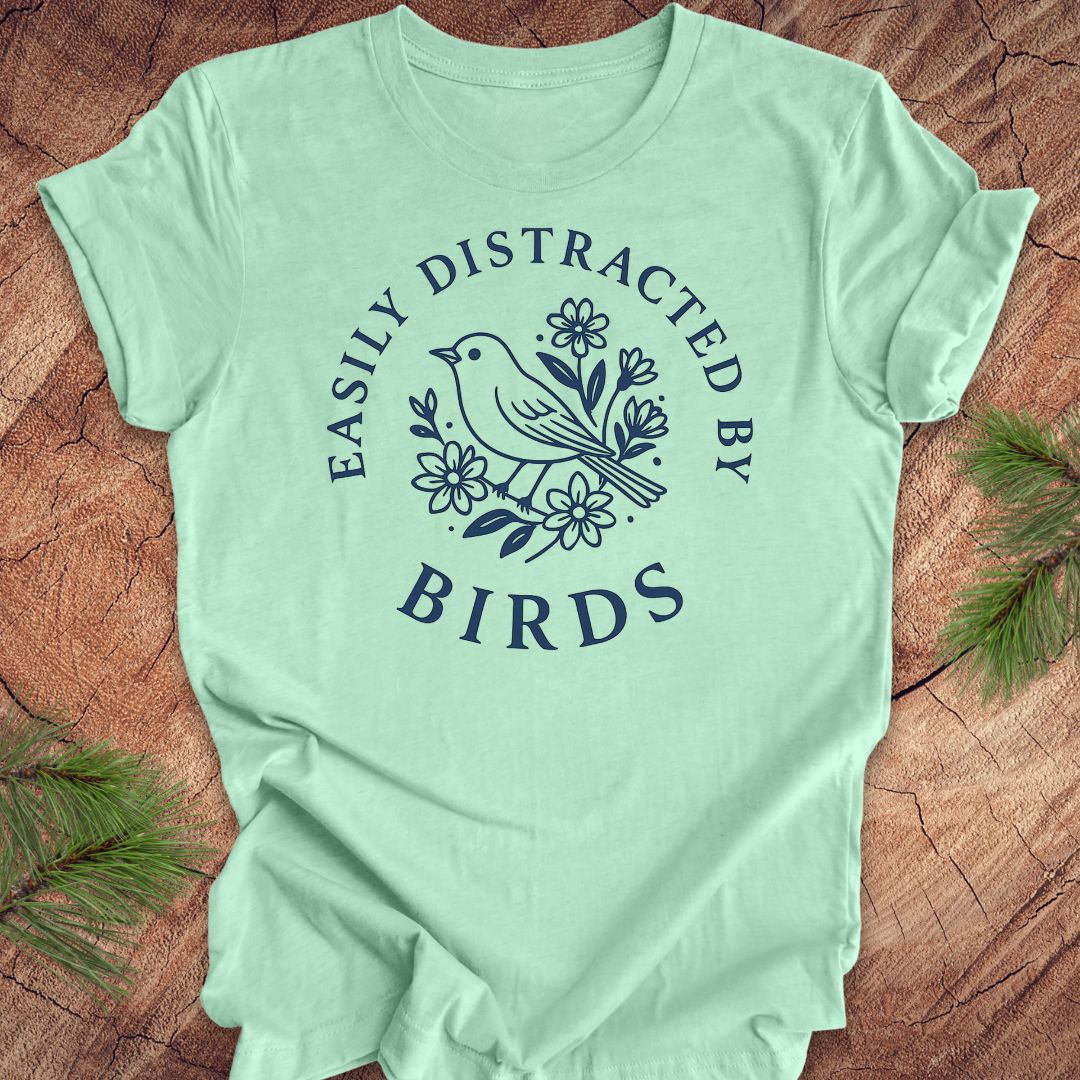 Mint green t-shirt with a bird graphic and text 'easily distracted by birds' on a wood background.