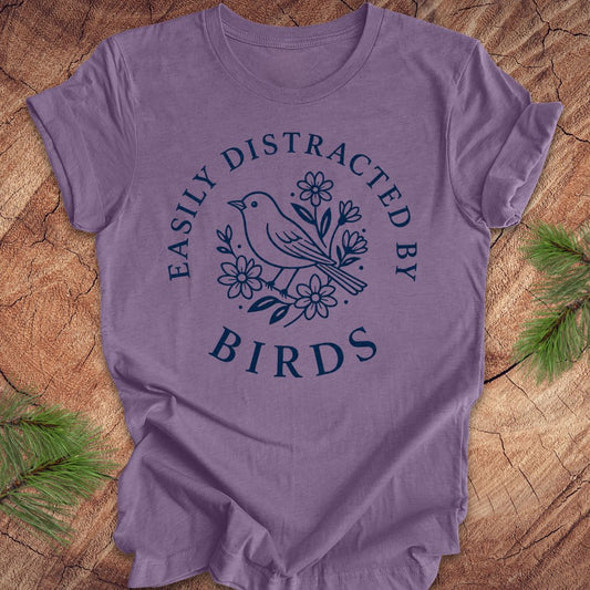 A lavender purple short-sleeve t-shirt with a navy blue graphic print of a bird and flowers, surrounded by the text 'EASILY DISTRACTED BY BIRDS'. The shirt is laid flat on a rustic wood background with pine branches in the corners.