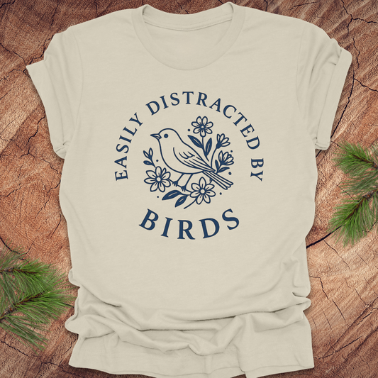 Beige t-shirt with blue graphic and text 'Easily Distracted by Birds' on a wooden surface.
