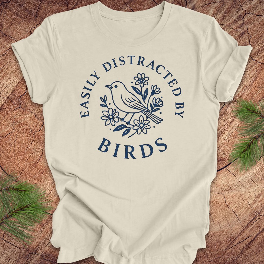Beige t-shirt with a bird graphic and text 'easily distracted by birds' on a wood background.