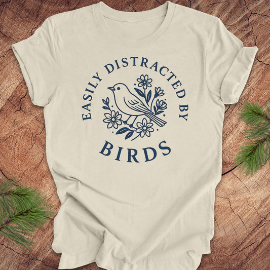 Beige t-shirt with a bird graphic and text 'easily distracted by birds' on a wood background.