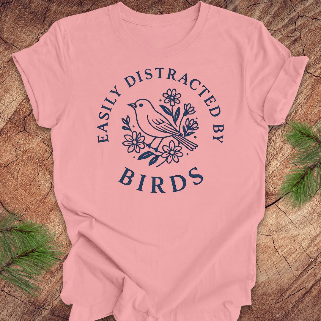 Pink t-shirt with a bird graphic and text 'easily distracted by birds' on a wood background.