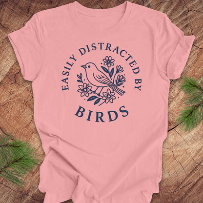 Pink t-shirt with a bird graphic and text 'easily distracted by birds' on a wood background.