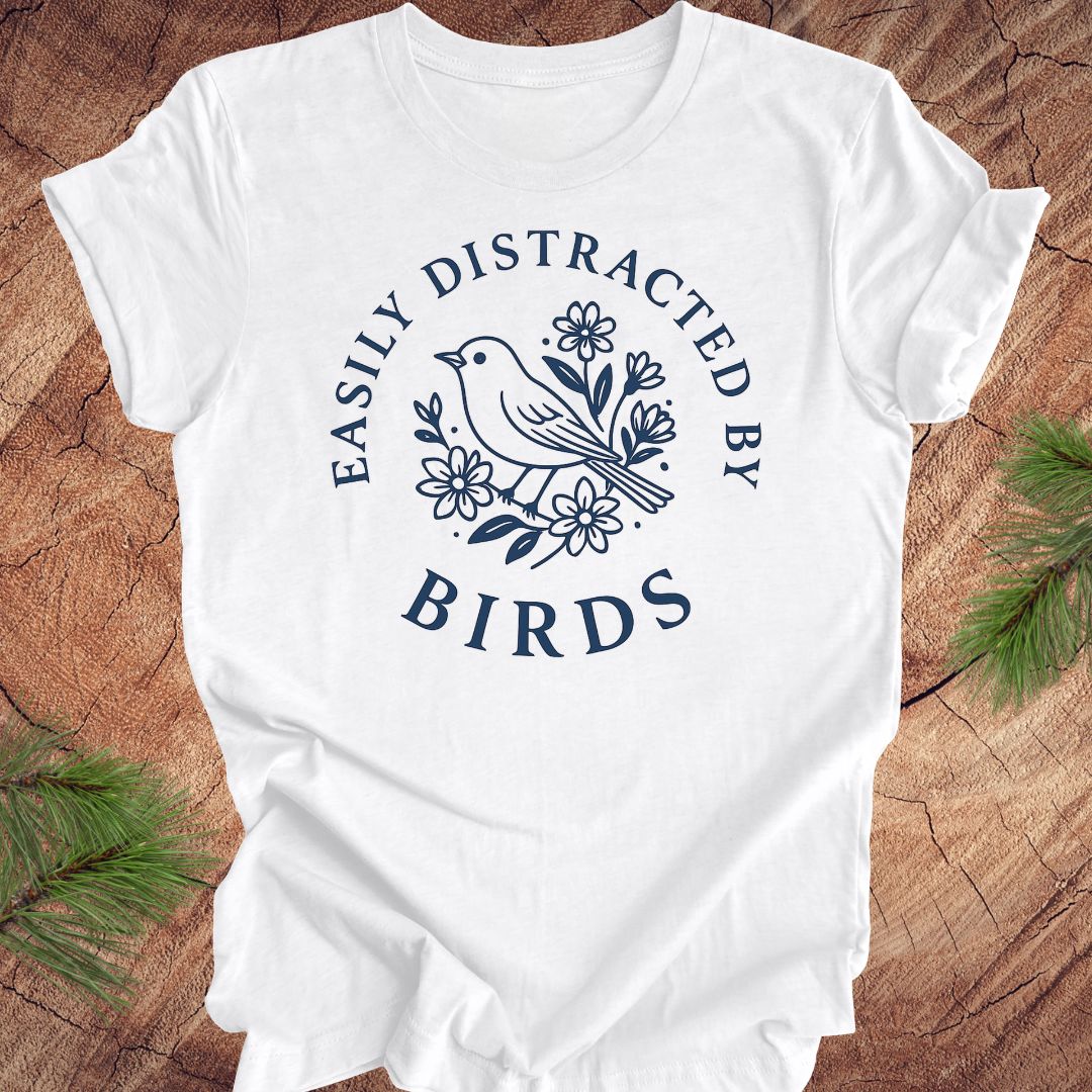 White t-shirt with a bird graphic and text 'easily distracted by birds' on a wood background.