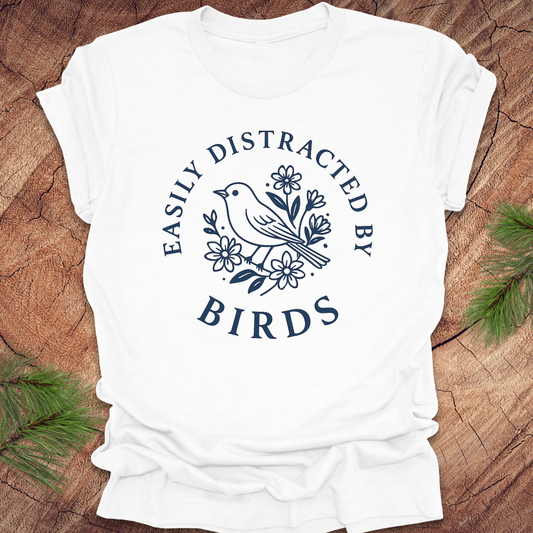 White t-shirt with a bird graphic and text on a wooden surface with greenery.