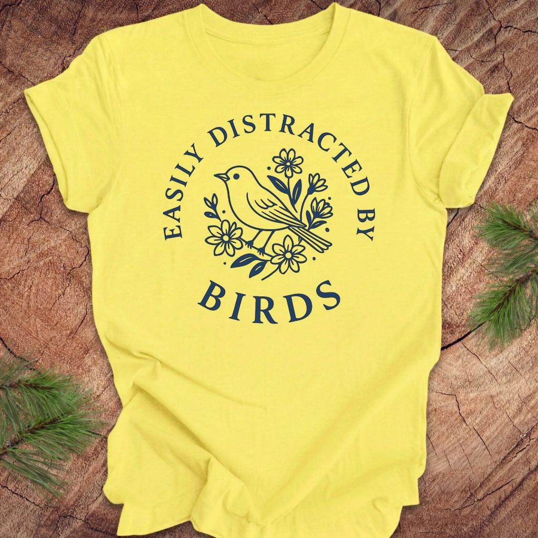 Yellow t-shirt with a bird graphic and text 'easily distracted by birds' on a wood background.