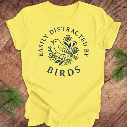 Yellow t-shirt with a bird graphic and text 'easily distracted by birds' on a wood background.
