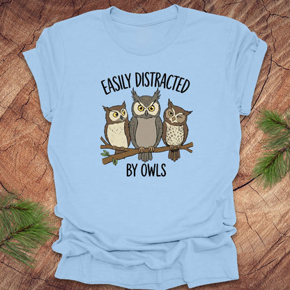 Baby blue t-shirt with owl design and text 'Easily Distracted by Owls' on a wooden surface.