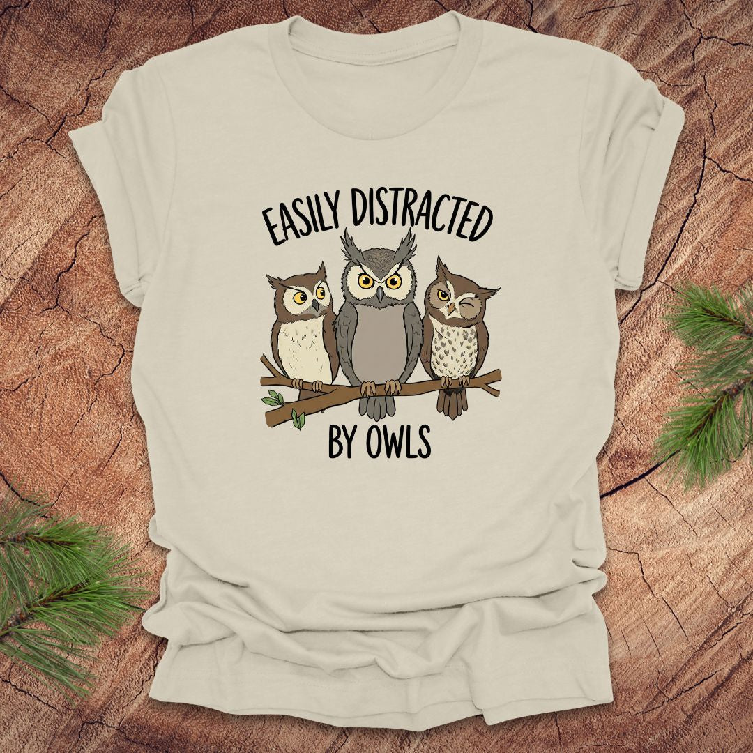 Beige t-shirt with owl design and text 'Easily Distracted by Owls' on a wooden surface.