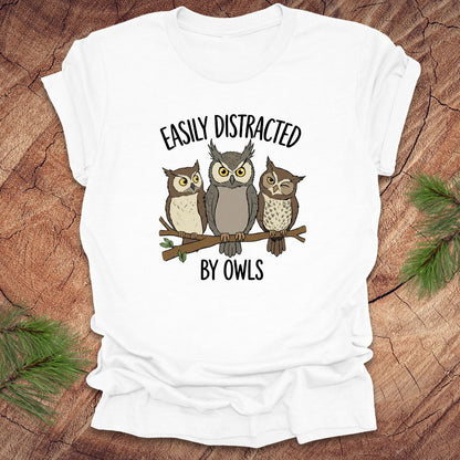 White t-shirt with owl design and text 'Easily Distracted by Owls' on a wooden surface.