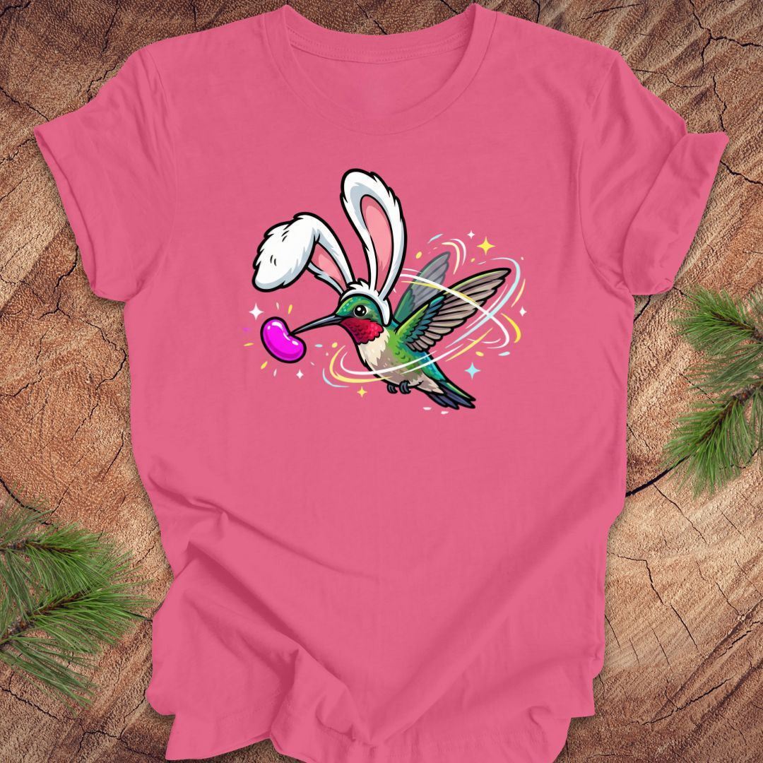 Pink t-shirt with a colorful graphic of a hummingbird with bunny ears on a wooden surface.