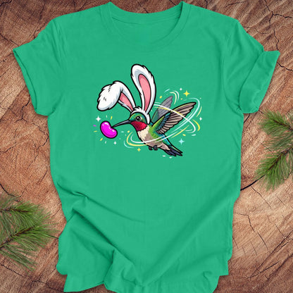 Green t-shirt with a cartoon design of a hummingbird wearing rabbit ears on a wooden surface.