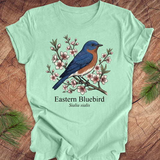 Light green t-shirt with an illustration of an Eastern Bluebird and floral design on a wooden surface.