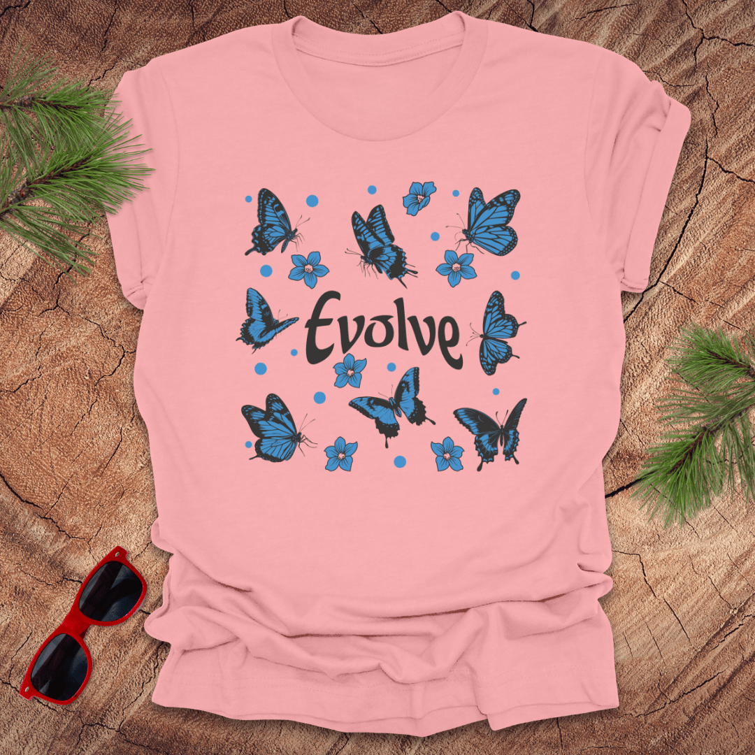 Pink t-shirt with blue butterflies and 'Evolve' text on a wooden surface with sunglasses and greenery.