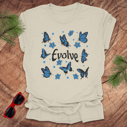 Beige t-shirt with blue butterfly design and 'Evolve' text on a wooden surface with sunglasses and greenery.