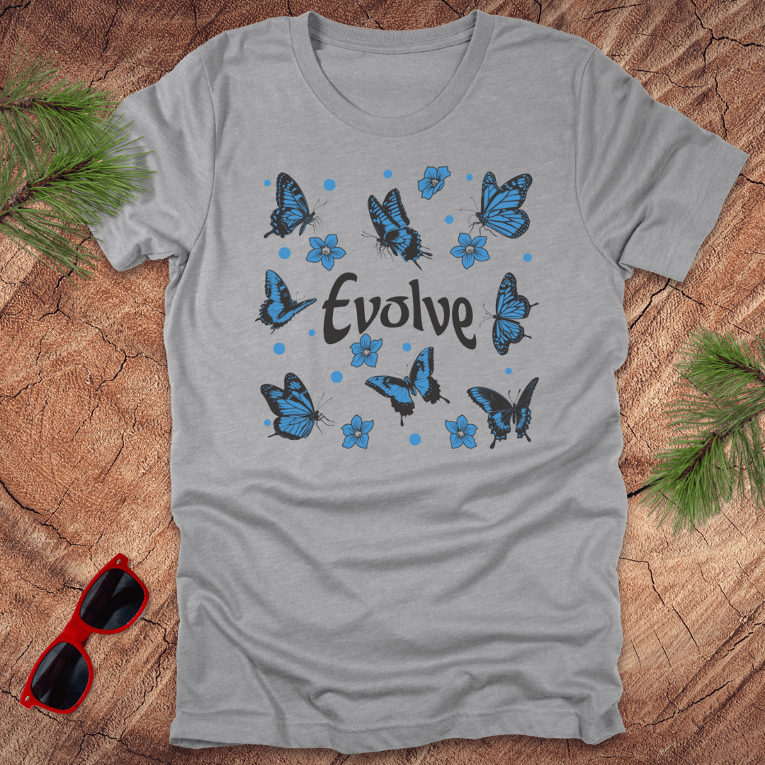 Gray t-shirt with blue butterflies and 'Evolve' text on a wooden surface with sunglasses and greenery.