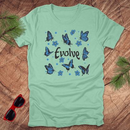Light green t-shirt with blue butterfly design and 'Evolve' text on a wooden surface with sunglasses and leaves.