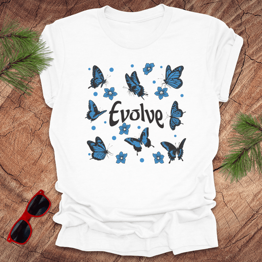 White t-shirt with blue butterfly design and 'Evolve' text on a wooden surface with sunglasses and greenery.