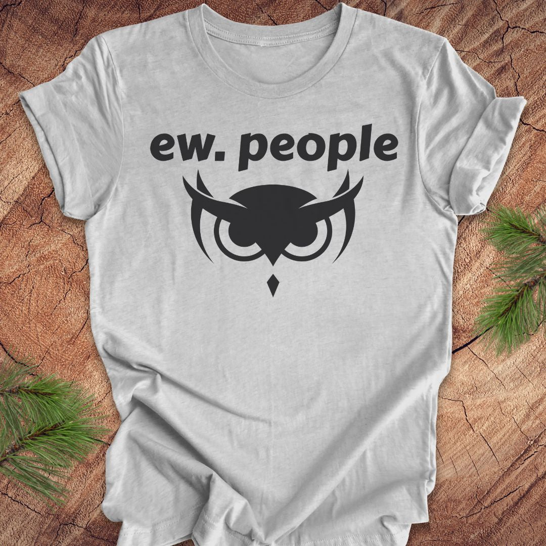 A light gray heather crewneck t-shirt with a black graphic print reading 'ew. people' above an owl face design, laid flat on a rustic wood background with pine branches in the corners.