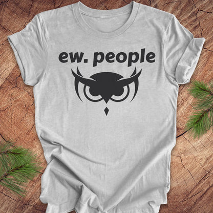 A light gray heather crewneck t-shirt with a black graphic print reading 'ew. people' above an owl face design, laid flat on a rustic wood background with pine branches in the corners.