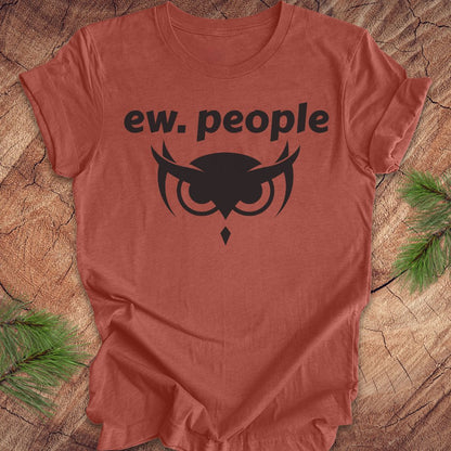 A terracotta-colored short-sleeve t-shirt with a black graphic print reading 'ew. people' above an owl illustration, laid flat on a rustic wood background with pine branches in the corners.