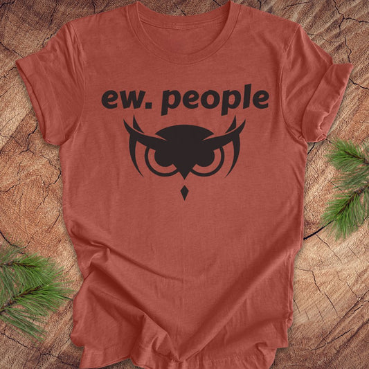 A terracotta-colored short-sleeve t-shirt with a black graphic print reading 'ew. people' above an owl illustration, laid flat on a rustic wood background with pine branches in the corners.